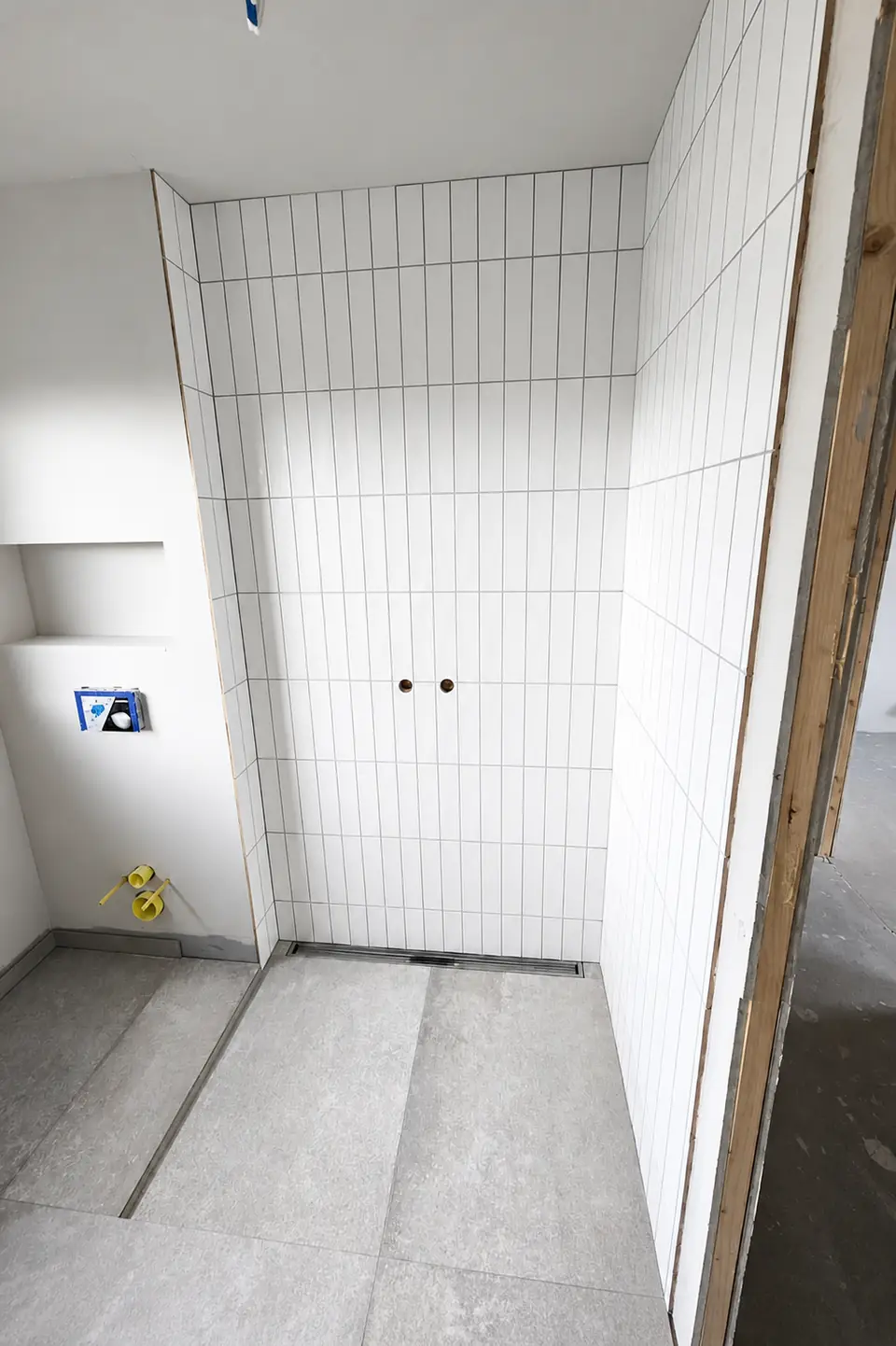 Bruseniche med hvide kvadratiske vægfliser i lige forbandt, grå gulvklinker og en linjeafløbsrende langs væggen