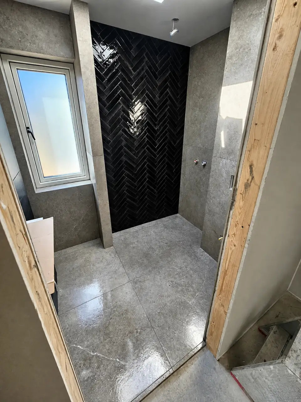 Badeværelse under opbygning med grå betonlook-fliser og en bruseniche i sorte blanke sildebensfliser, set mellem uafsluttede trækarme